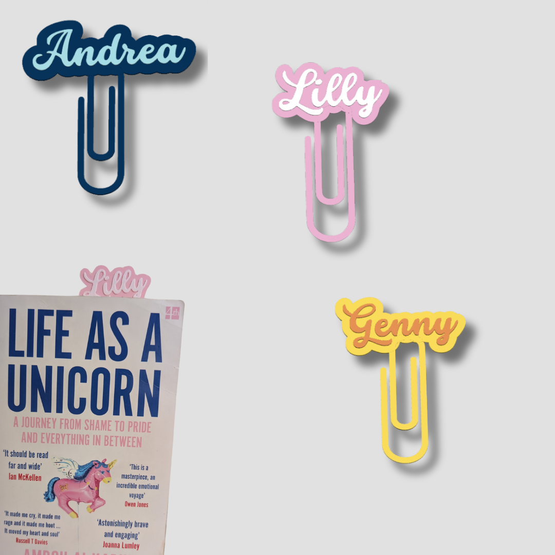 Group of three personalised paperclip bookmarks in blue ('Andrea'), pink ('Lilly'), and yellow ('Genny'), showing the clips on a book.