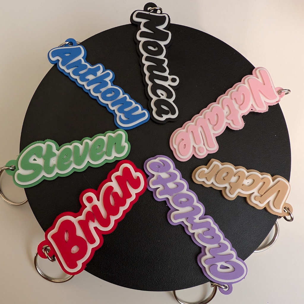 Group of custom name keyrings in bright colours (red, blue, green, pink, purple) with names Anthony, Monica, Steven, and Brian, displayed on a black background.
