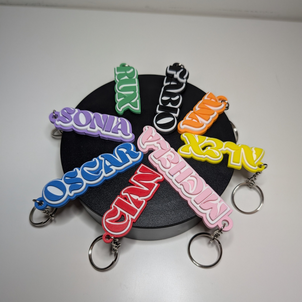 Group of custom name keyrings in bright colours (purple, blue, red, pink, green, yellow) with names like Oscar, Ruby, and Fabio, displayed on a black background.